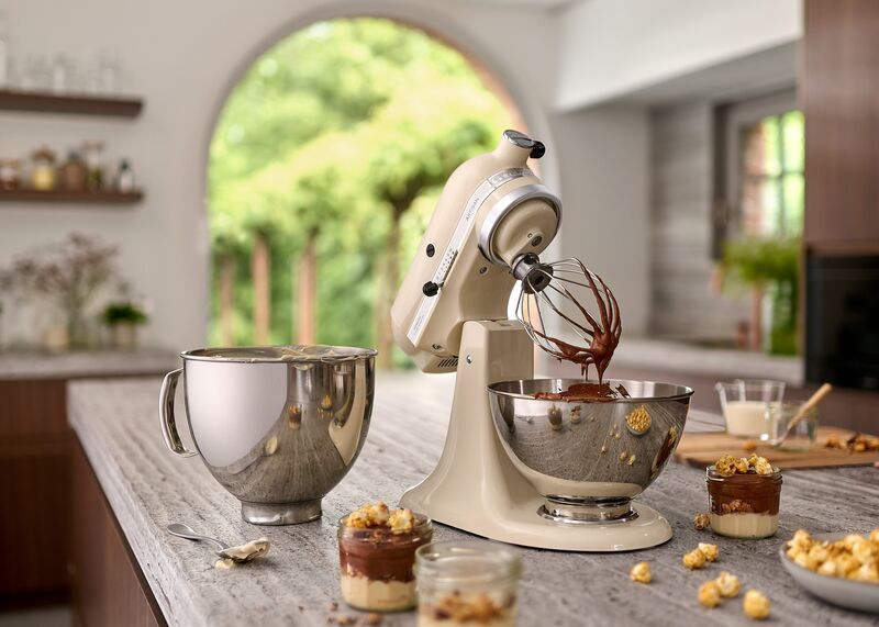 KitchenAid mikser miješa čokoladnu smjesu u zdjeli, s dodatnom posudom i desertima u pozadini na kuhinjskoj radnoj površini.