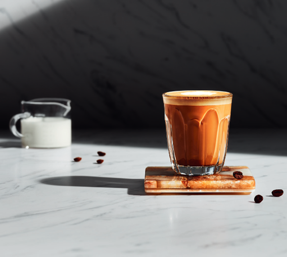 Staklena čaša s cortado kavom na mramornoj površini, uz mlijeko i zrna kave u pozadini.