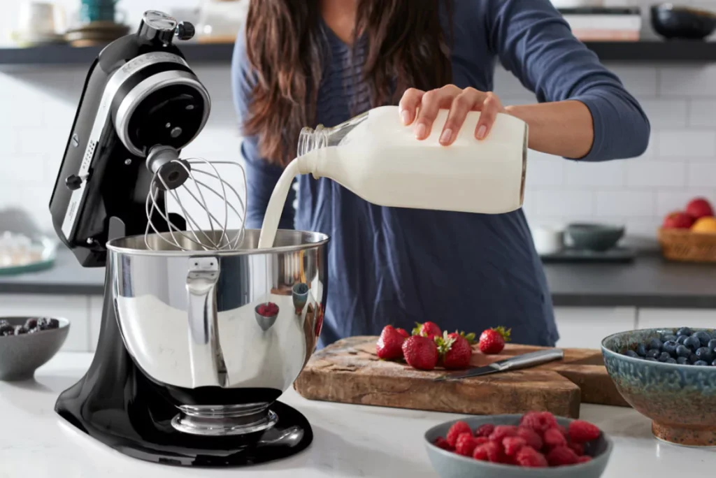 Žena ulijeva mlijeko u KitchenAid stolni mikser u modernoj kuhinji, okružena svježim bobičastim voćem.